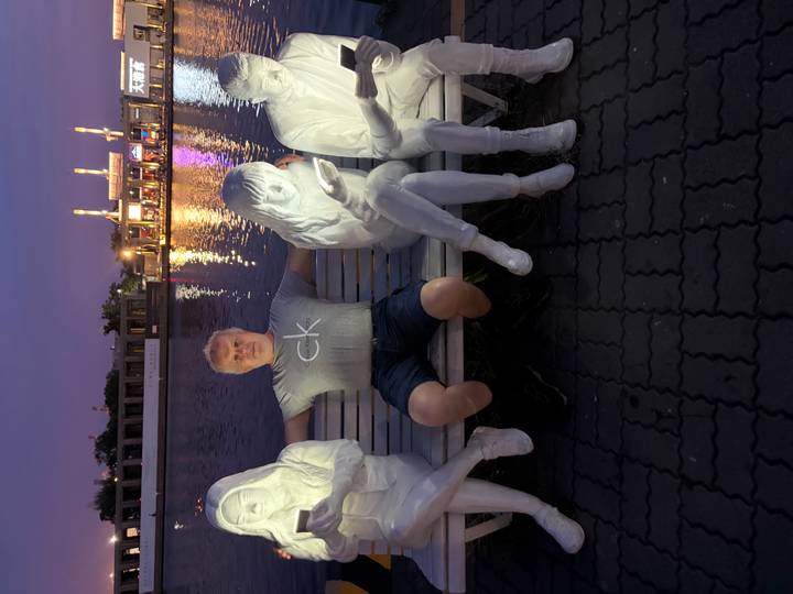 A man poses on a bench flanked by illuminated white statues beside calm evening water.