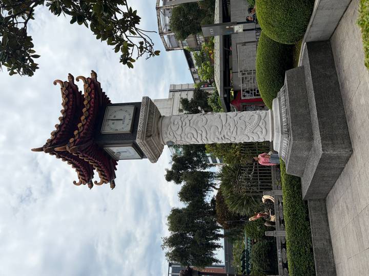 A traditional pagoda-style clock tower mounted on a decorative stone pedestal in a small park.