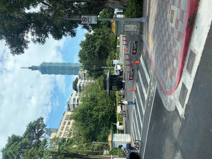 Street scene with Taipei 101 rising above trees and buildings beyond a crosswalk and traffic cones.