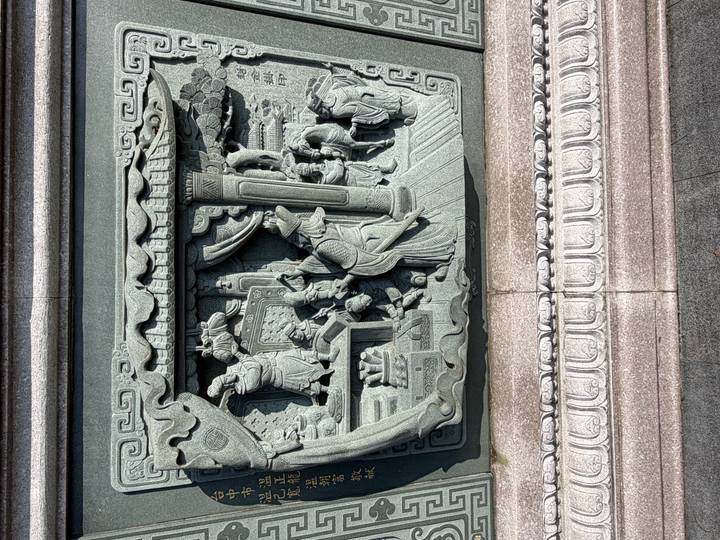 Sun-lit relief carving on green stone portraying an intricate domestic scene with figures and furnishings.