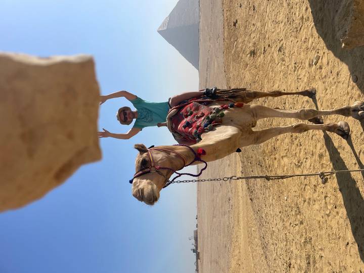 Traveler with raised arms riding a camel across the sandy plateau near the pyramids on a sunny day.