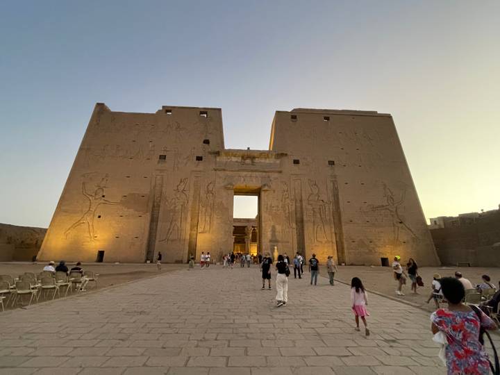 Visitors walk toward the illuminated sandstone entrance of Edfu Temple at dusk.