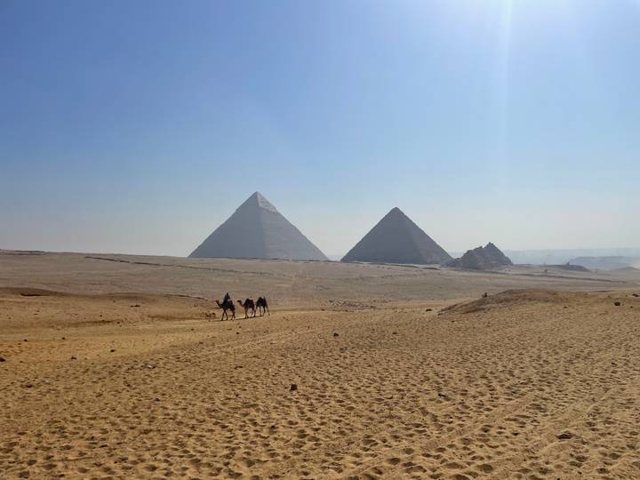 Wide desert vista with two main pyramids on the horizon and a small caravan of camels crossing the sands.