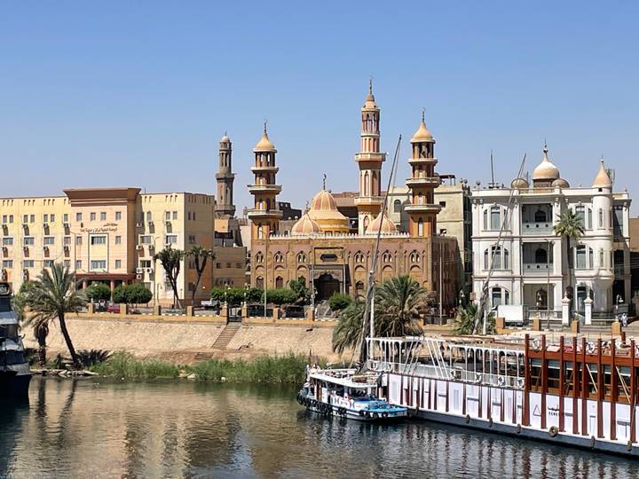 Daytime riverside cityscape featuring a riverside mosque, colonial buildings, and moored cruise boats on the Nile.