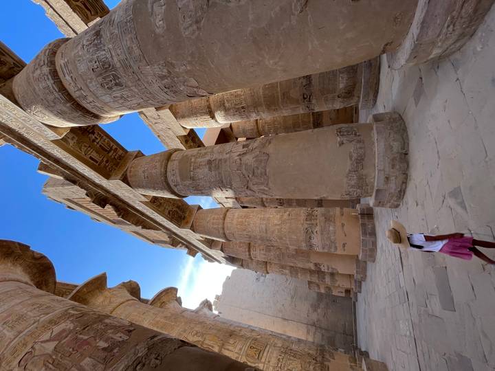 Young girl in a hat walks beneath towering sandstone columns bathed in afternoon light inside Karnak Temple.