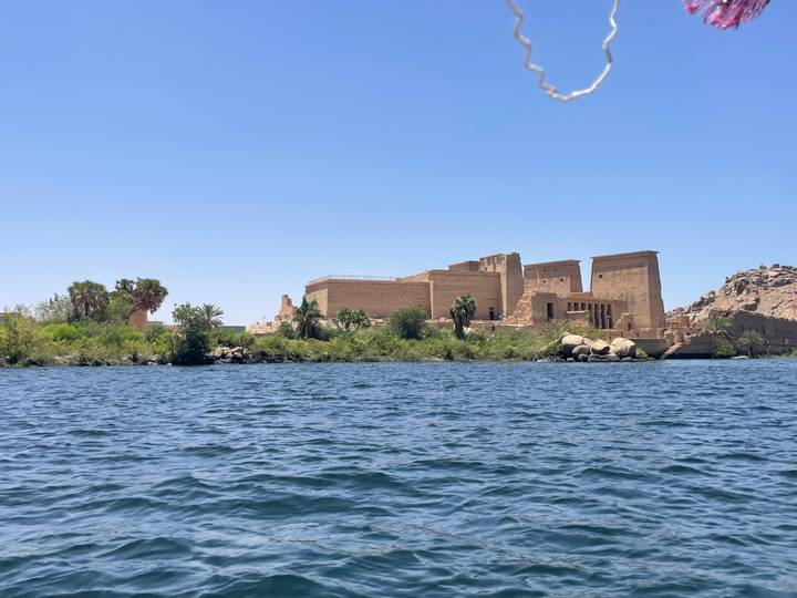 Philae Temple complex seen from a boat on the shimmering blue Nile with greenery along the shoreline.