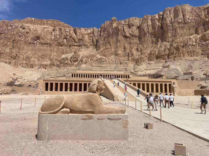 Temple of Hatshepsut nestled against dramatic cliffs with visitors exploring the monumental ramp and sphinx statue in foreground.