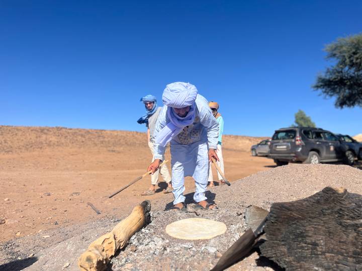 Local guide in traditional blue attire prepares bread over coals while travelers watch in desert setting.