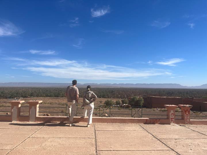 Two travelers pause at a scenic overlook with expansive palm groves and distant mountains beneath a blue sky.