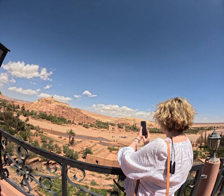 Woman taking a photo of the mud-brick fortified village stretching across a desert valley under a bright blue sky.