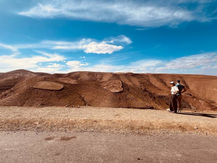 Two people pause on a roadside overlooking rolling, barren desert hills beneath a vivid blue sky with scattered clouds.