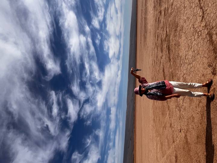 A woman stands on cracked desert earth beneath dramatic swirling clouds, pretending to pinch the distant horizon.