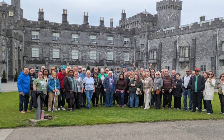 Large tour group poses on the lawn in front of a gray-stone castle with battlements and tall chimneys.