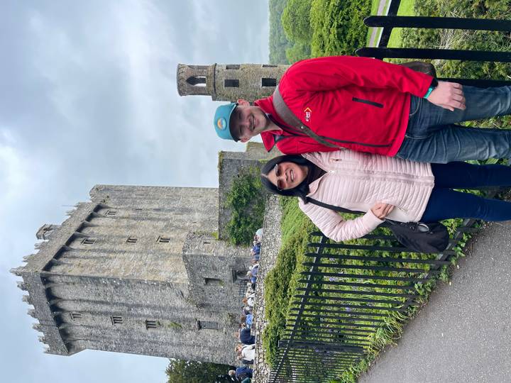 Two travelers smile for a photo outside a tall medieval stone castle surrounded by lush greenery.