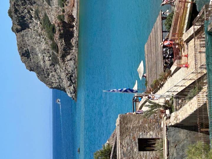 Turquoise bay with stone buildings, a Greek flag, and parasols beside a steep rocky headland.