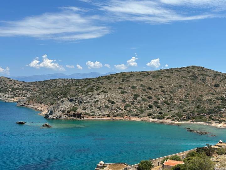 Rocky peninsula and clear turquoise sea with distant mountains under a blue sky.
