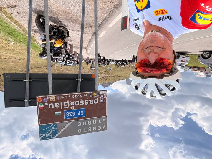 Cyclist taking selfie at mountainous Passo Giau road sign with motorbikes nearby