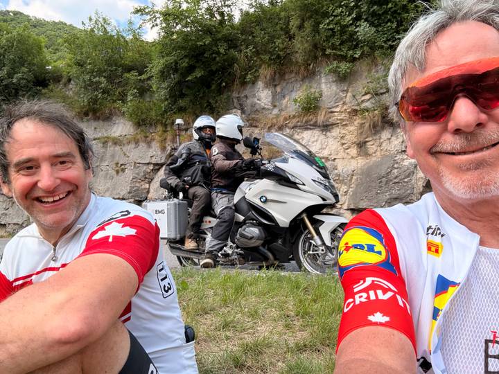Cyclists chatting roadside as motorcycle camera crew passes in background