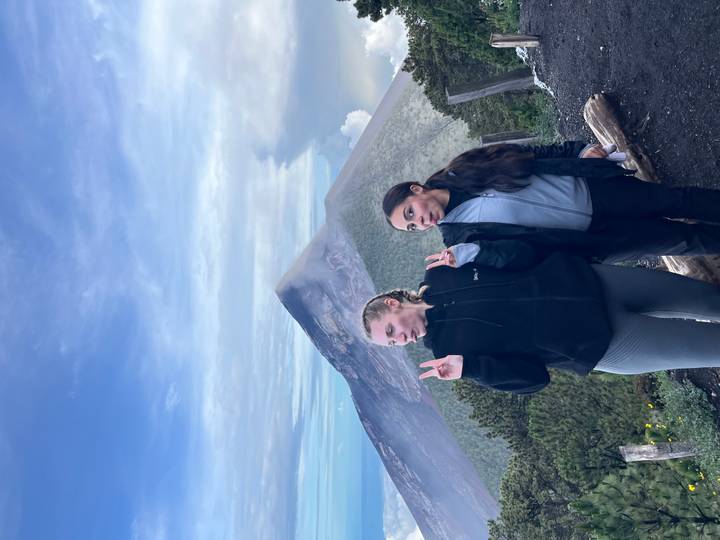 Two young women posing with peace signs on a mountaintop with a smoking volcano peak behind them
