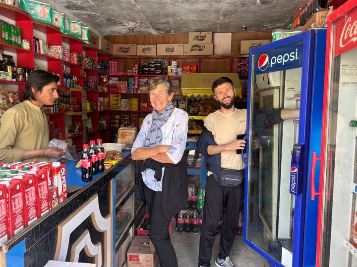 Two travellers chat with a local shopkeeper inside a colourful convenience store stocked with snacks and drinks.