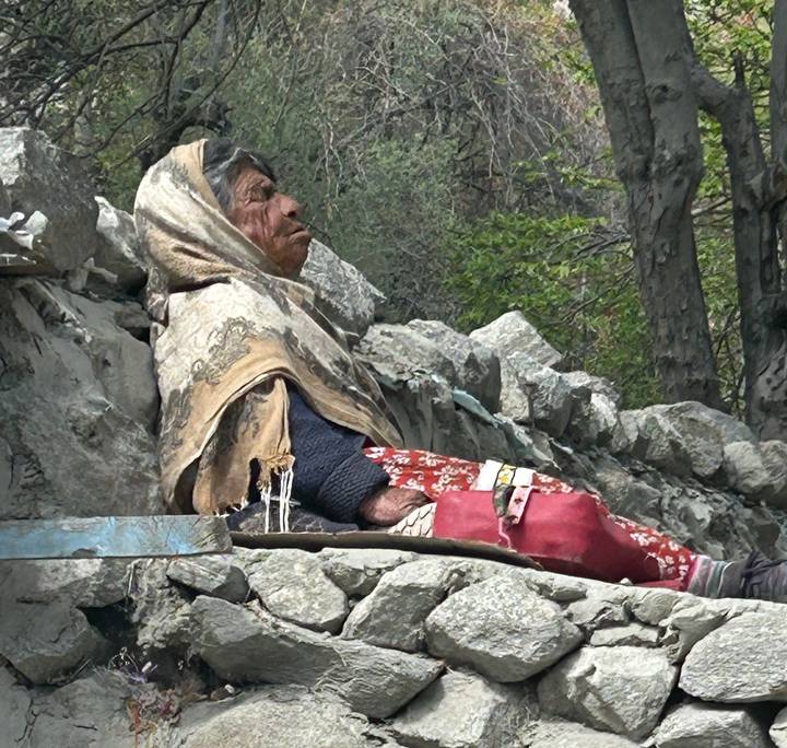 A weather-worn, shawl-covered figure seated amongst rough stone blocks in an outdoor setting.