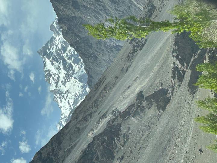 Steep rocky slope leading up to a striking snow-covered mountain peak beneath scattered clouds.