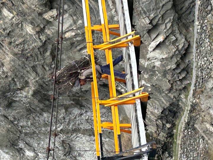 Local man carrying a heavy bundle of sticks across a narrow yellow suspension bridge over rocky terrain.