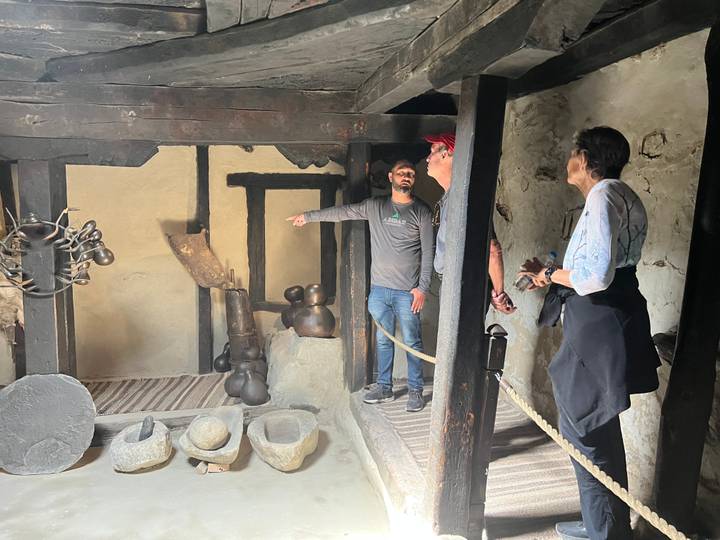 Guide points out historic household items to two travellers inside a dimly lit traditional room.