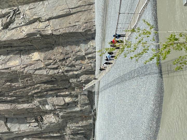 Travellers carefully cross a long cable bridge suspended beside towering rock cliffs.