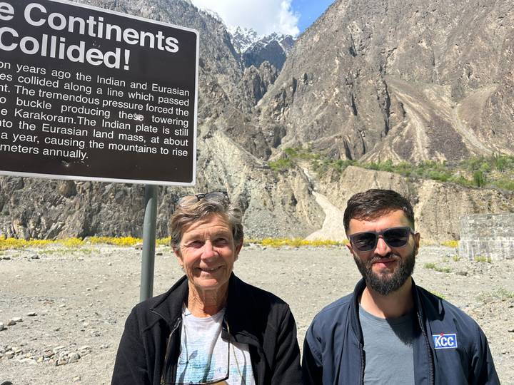 Two travellers stand by an informational geology sign with steep rocky mountains behind.