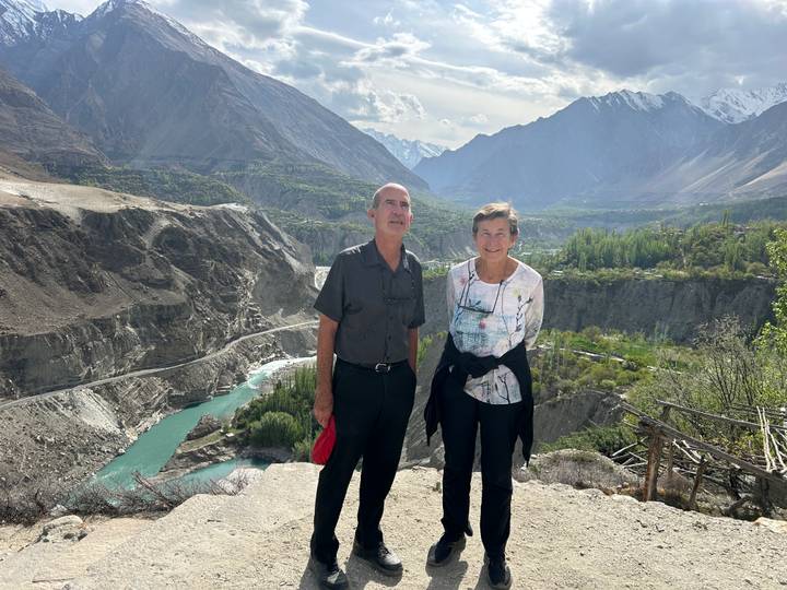 Travellers pose at a viewpoint over a turquoise river winding through a steep canyon with snowy peaks beyond.