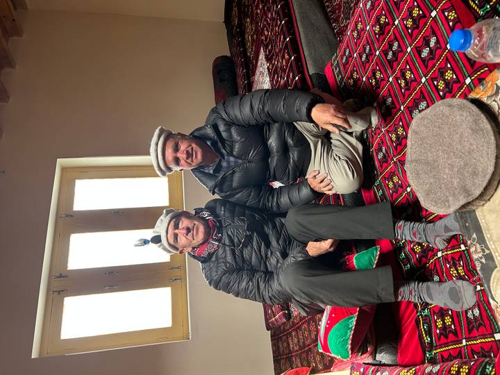 Two men wearing local caps sit on patterned carpets in a cosy, sunlit room.