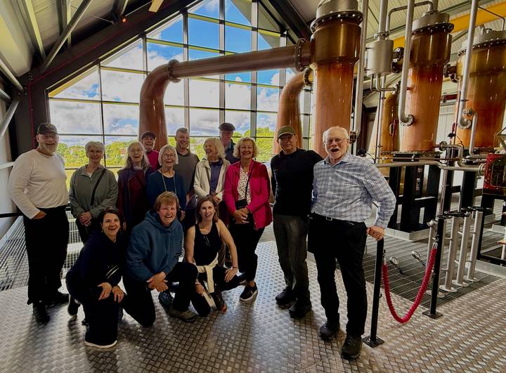 Large tour group posing inside a whiskey distillery with copper stills and big windows behind.