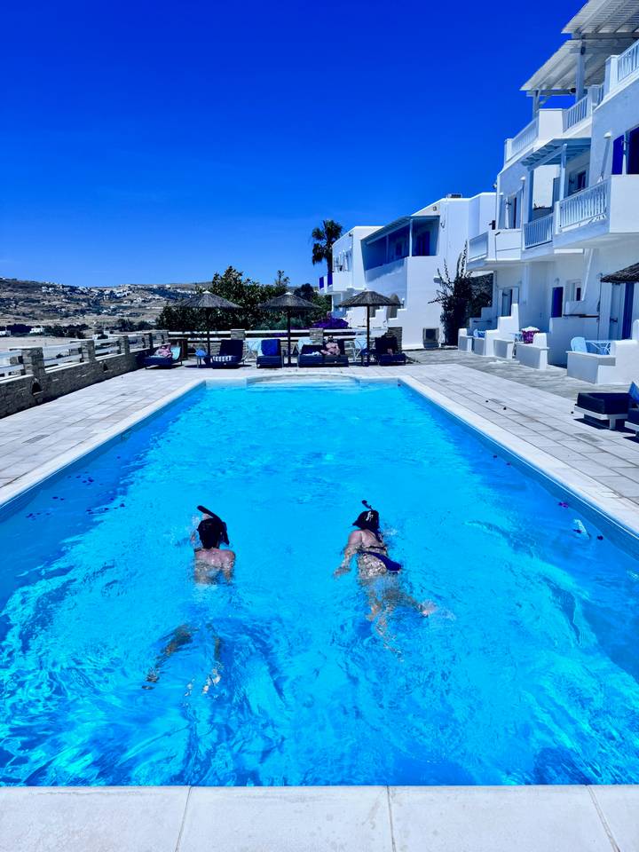 Two snorkellers glide across a bright blue hotel pool surrounded by sun-loungers and white Cycladic buildings.