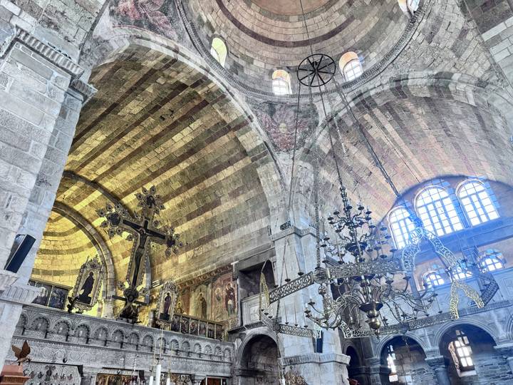 Interior view of a grand stone church with striped vaulted ceiling, chandeliers and religious icons.