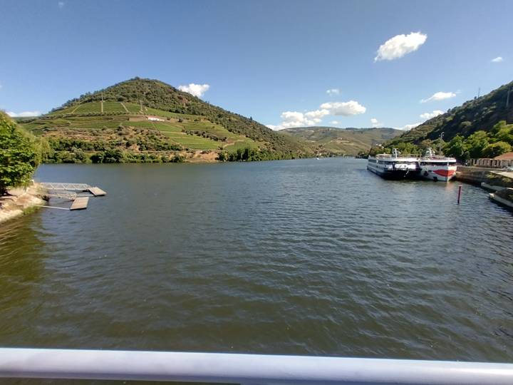 Wide Douro River flanked by terraced hillsides with two river-cruise boats docked under a bright sky.