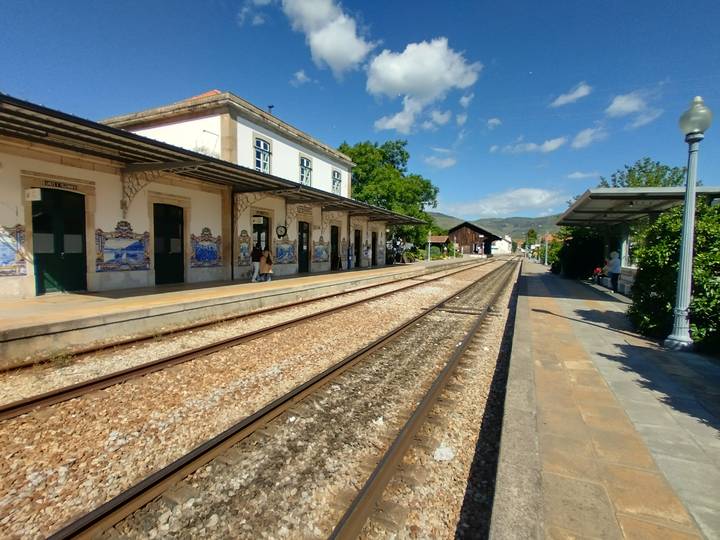 Historic railway station with azulejo tile panels, passengers waiting along sunlit tracks toward distant mountains.