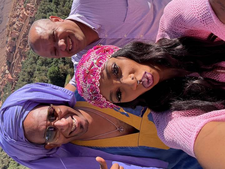 Three smiling travelers in vibrant headwear take a selfie with a lush Moroccan valley behind.
