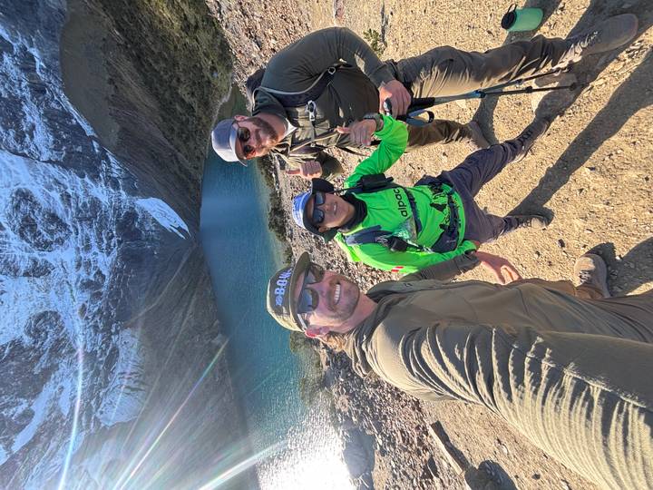 Three hikers posing beside turquoise alpine lake beneath a glacier wall