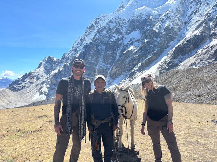 Travellers and a local horse handler posing with a white horse beneath rugged Andean cliffs