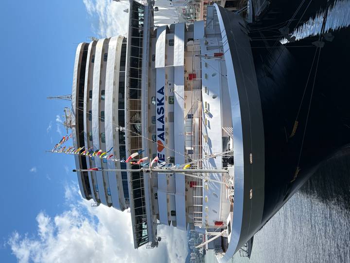 Large Holland America cruise ship docked with a banner reading 'I ♥ ALASKA' under blue sky.