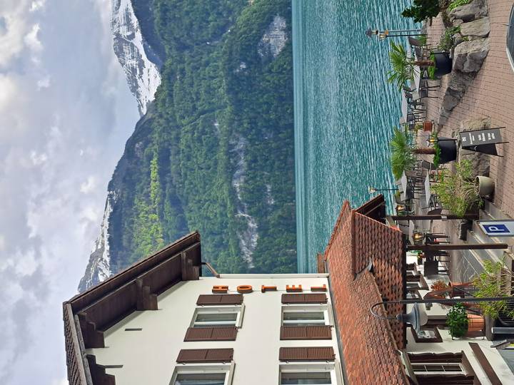 Lakefront alpine hotel with outdoor terrace overlooking turquoise water and snow-capped peaks under a lightly clouded sky.