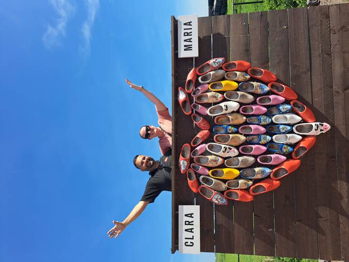 Smiling pair posing with arms raised behind a large heart made of colourful Dutch wooden clogs against a clear blue sky.