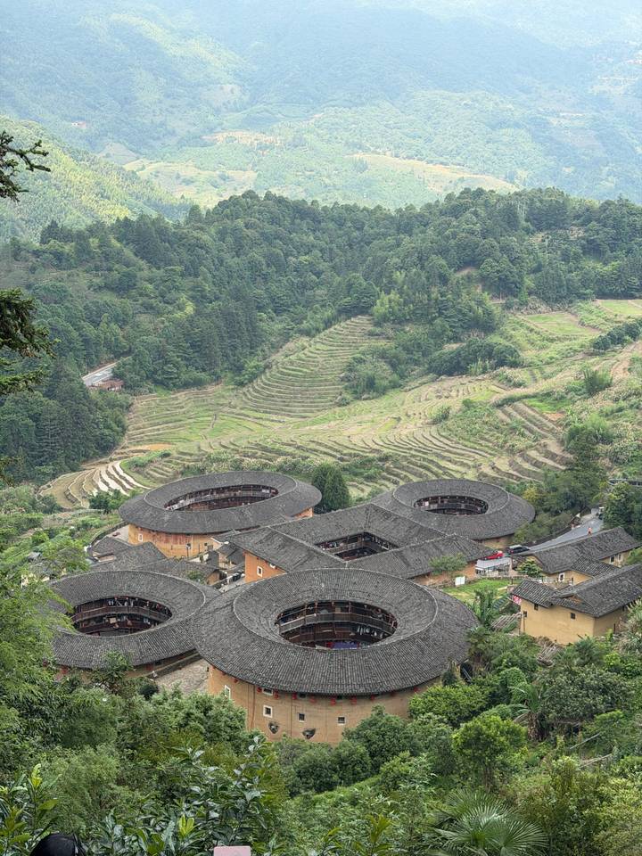 Aerial view of circular Hakka Tulou earth buildings amid terraced hills and pine forests.