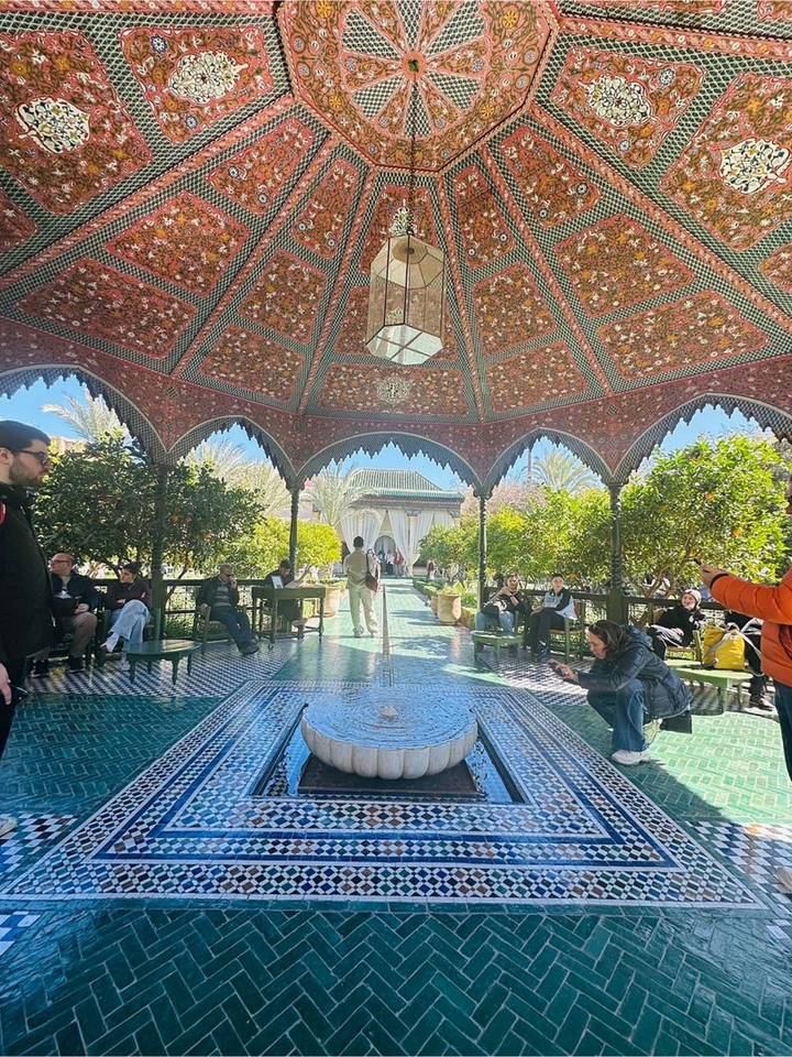 Visitors relax under an ornate tiled pavilion surrounded by orange trees and patterned mosaic flooring in a palace garden.