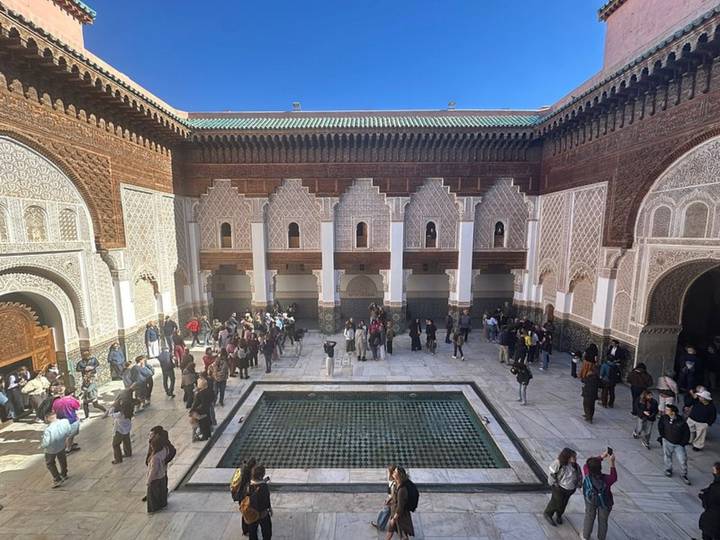 Wide view of the Ben Youssef Madrasa courtyard with visitors surrounding the central tiled pool and carved stucco façades.