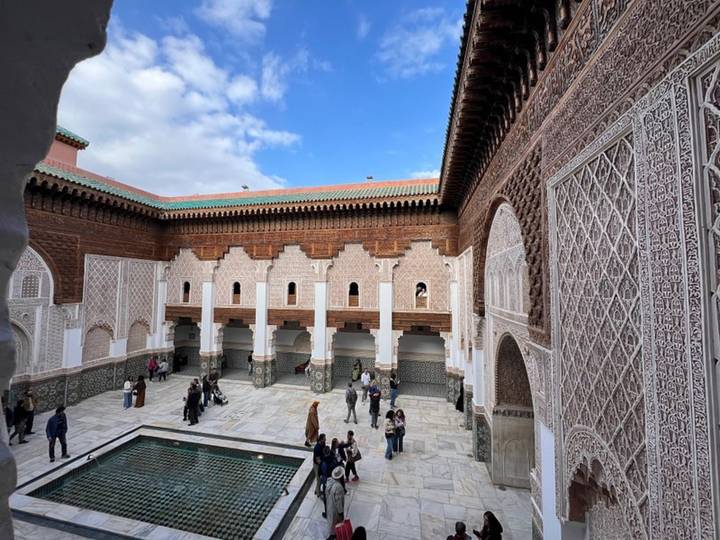 Angled view across the ornate courtyard of Ben Youssef Madrasa with tiled pool, carved arches and visiting tourists.