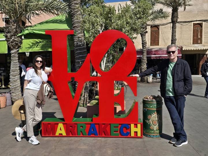 Smiling couple poses beside a large red LOVE Marrakesh sign on a palm-lined plaza.