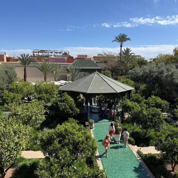 Lush courtyard garden with hexagonal gazebo shaded by orange trees, visitors strolling on green tile paths.