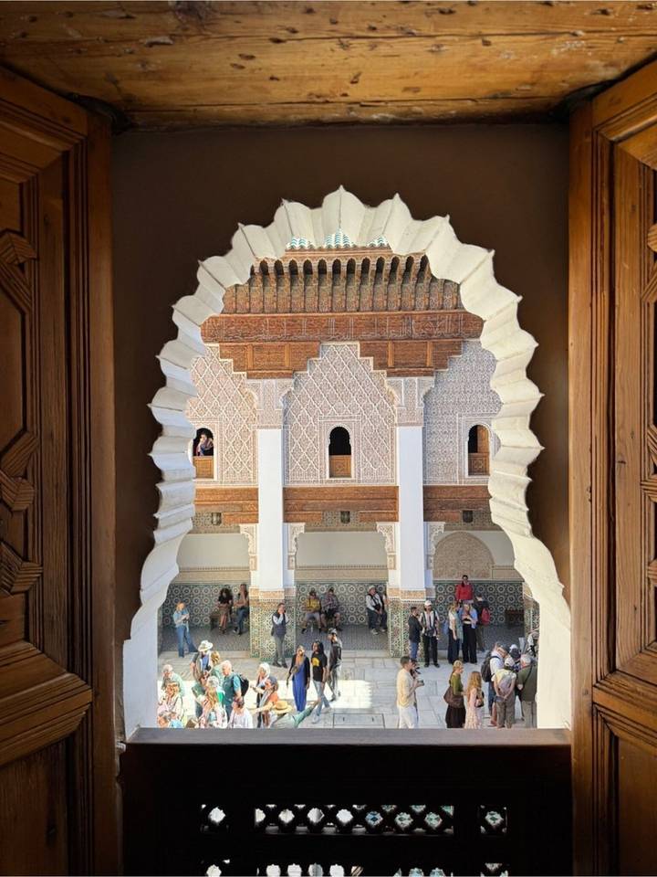 View through a sculpted keyhole window framing the courtyard and arches of a historic madrasa with visitors below.